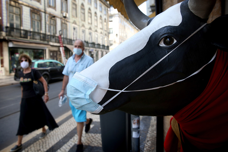 People wearing face masks pass by a statue of cow on a street in Lisbon